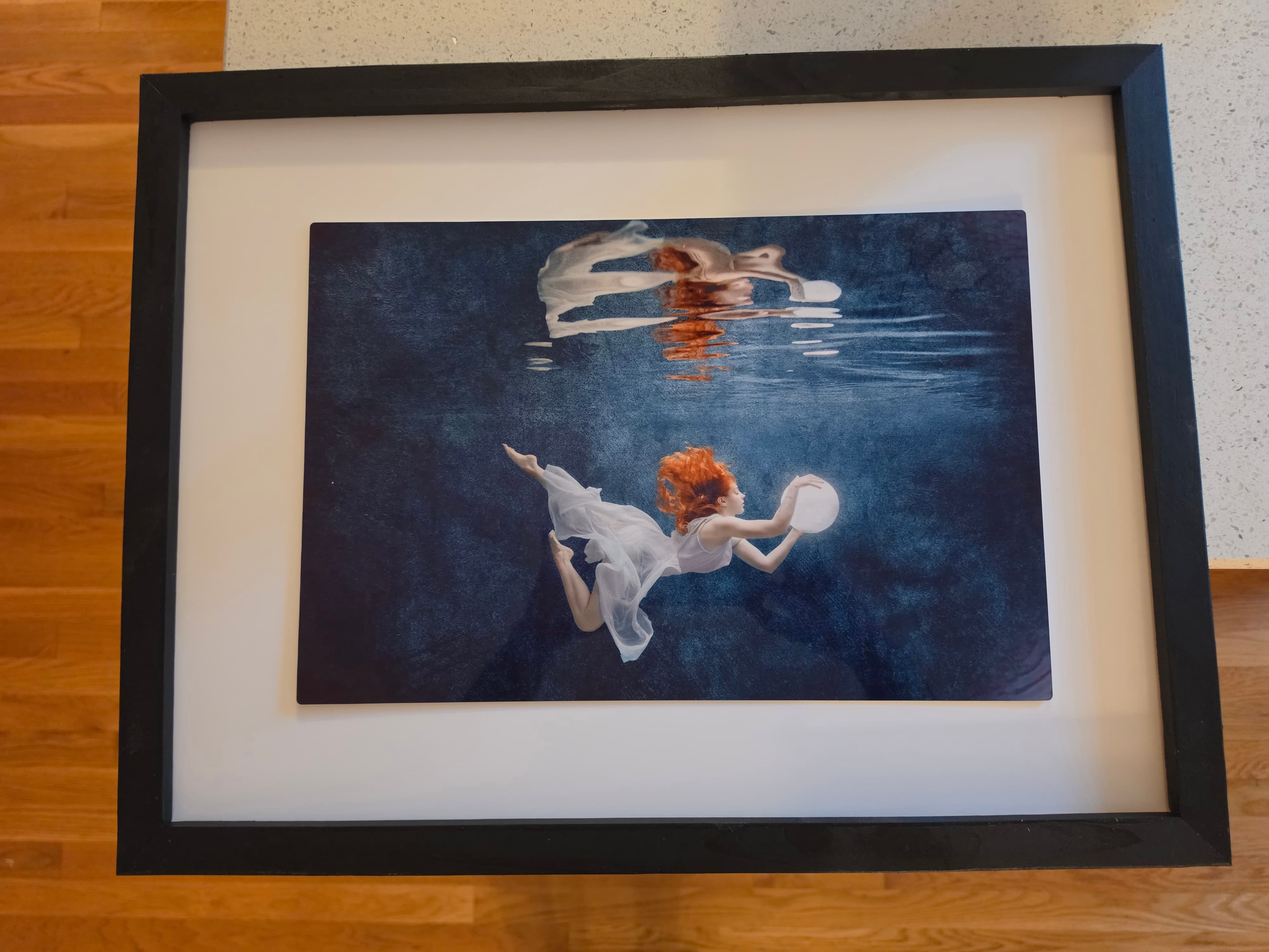 Framed photograph of a red-haired woman underwater in a white dress, holding a glowing white orb with a reflection shimmering at the water's surface.